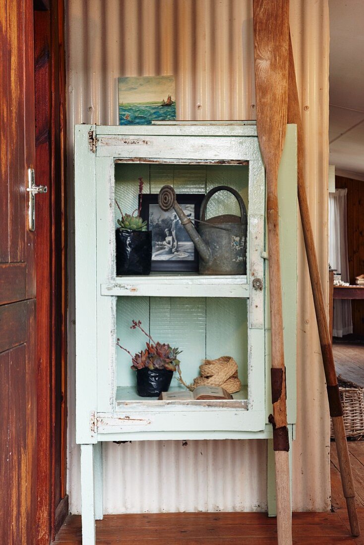 Mit Zinkgießkanne und Sukkulenten dekorierter Vintage Vitrinenschrank und zwei Holzpaddel vor blechverkleideter Wand