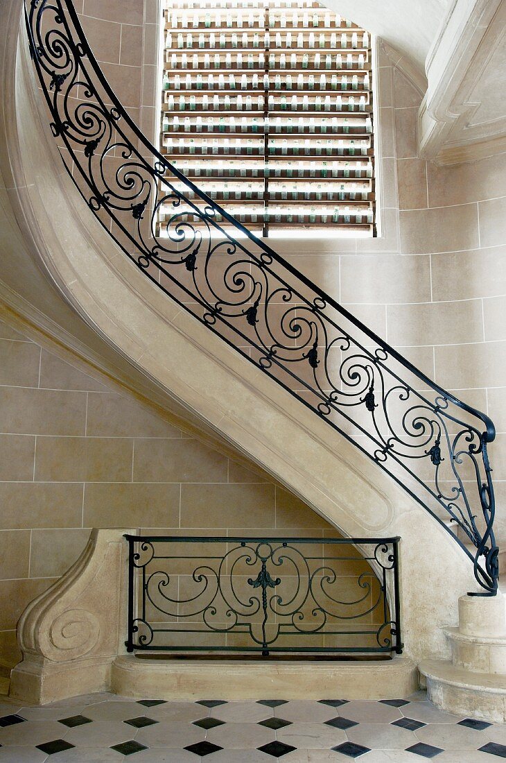 Curved staircase with wrought iron balustrade below window with closed blind in foyer with stone tiles on walls