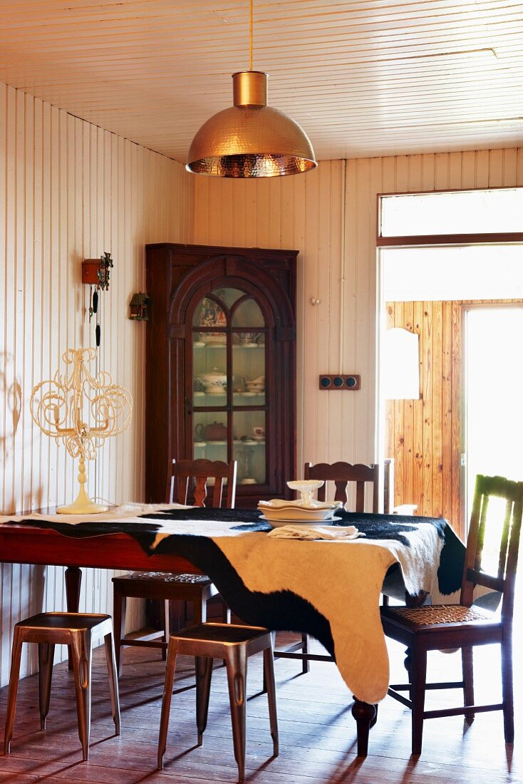 Antikstühle und Metallhocker an Esstisch mit Kuhfelldecke; Vitrineneckschrank vor holzvertäfelter Wand