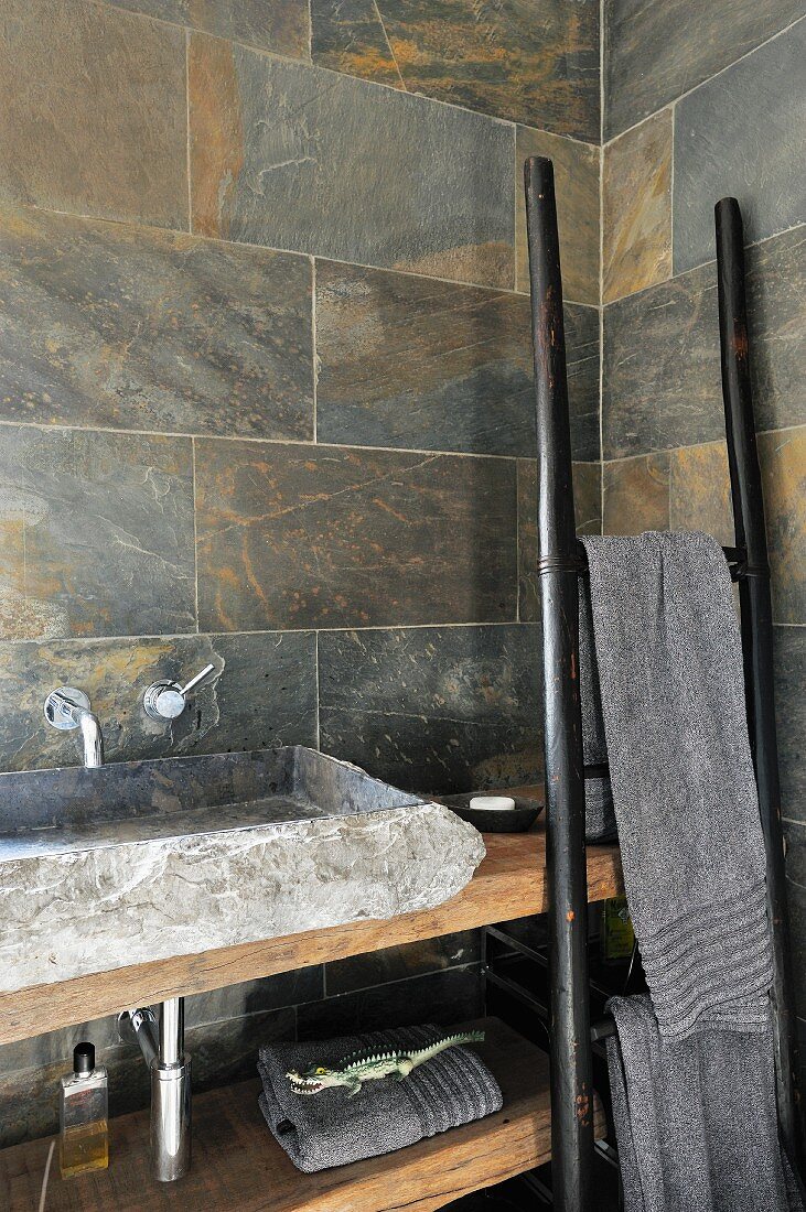 Rustikales Waschbecken aus Stein auf Holzplatte und angelehnter, leiterartiger Handtuchhalter in Badezimmerecke mit marmorierten Steinfliesen