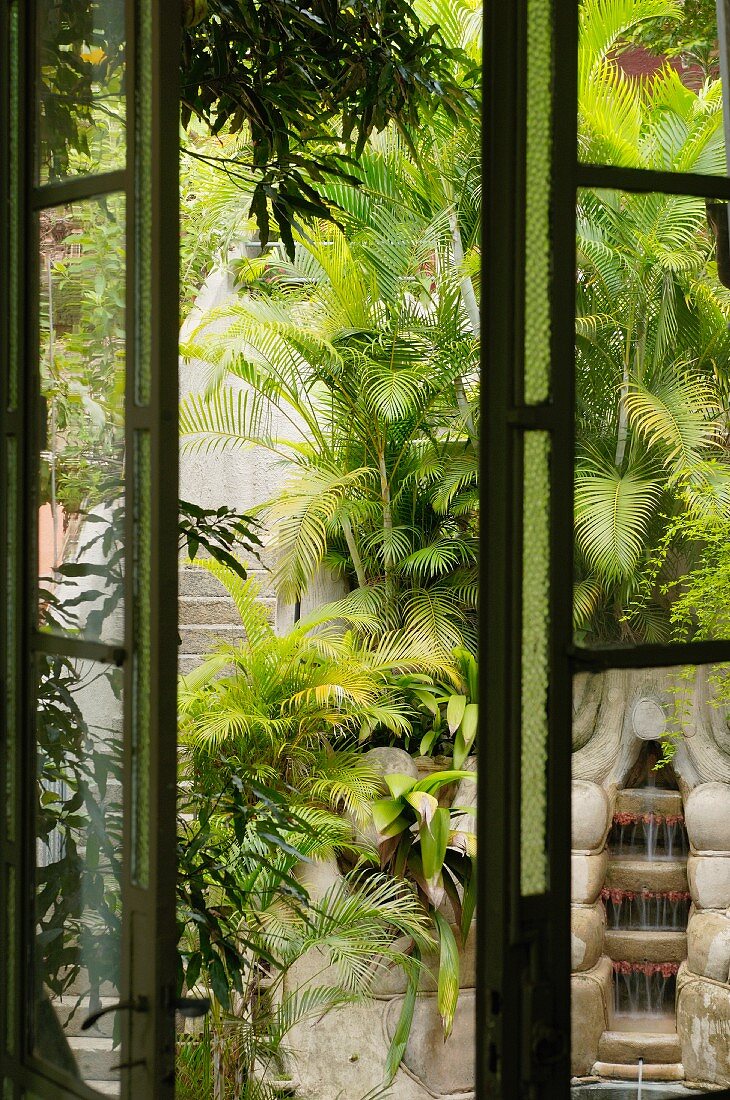 Blick durch halb geöffnetes Fenster auf palmenbewachsene Steintreppe und Kaskadenbrunnen