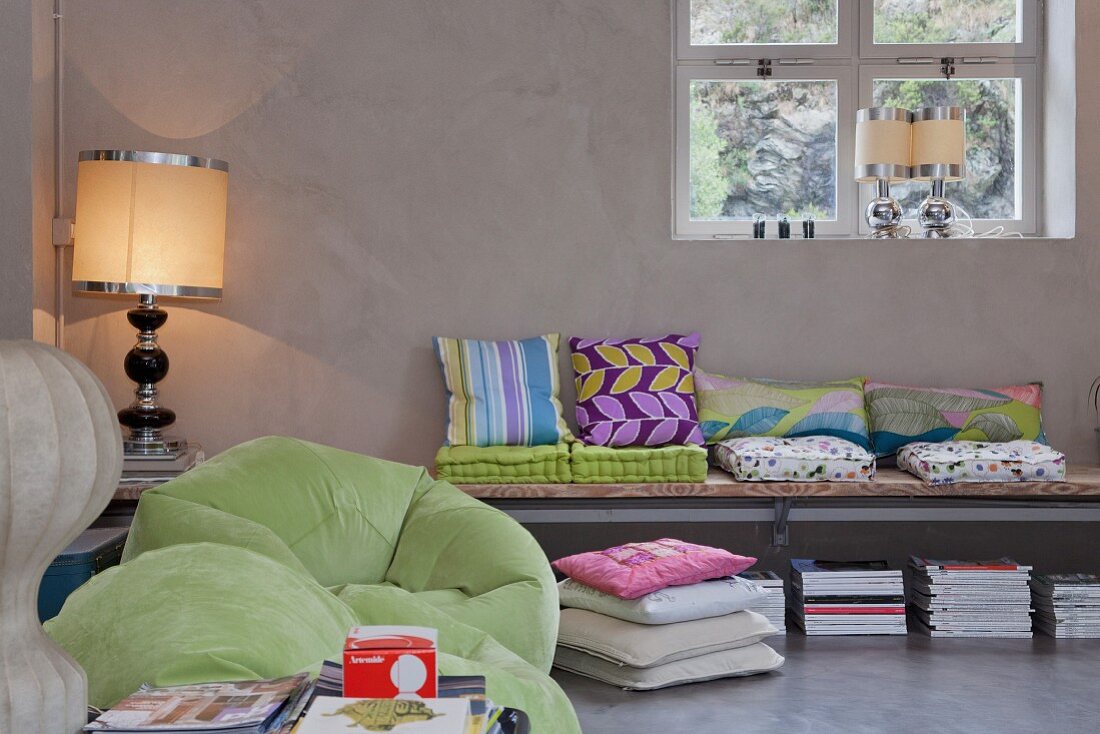 Collection of colourful cushions on wall-mounted bench, lime green beanbag chairs and postmodern table lamps contrasting with cool exposed concrete of loft interior