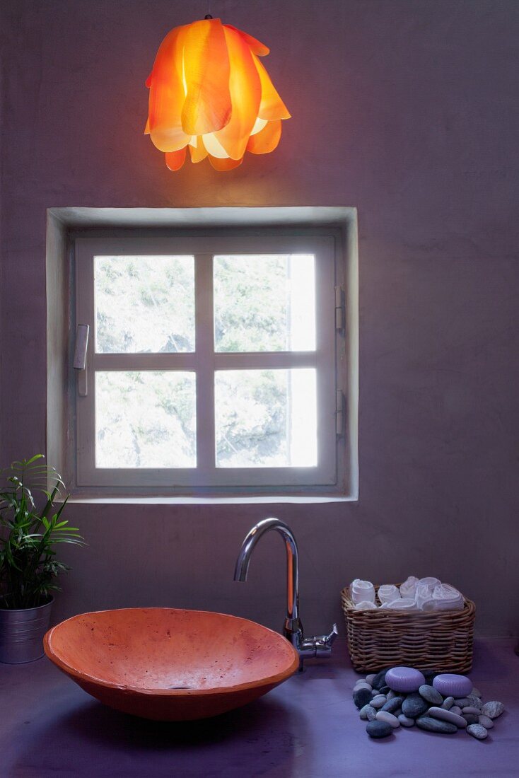 Stimmungsvolles Badinterieur mit Keramikschüssel und Hängeleuchte in warmem Orange zu pastellviolettem Waschtisch vor Sichtbetonwand