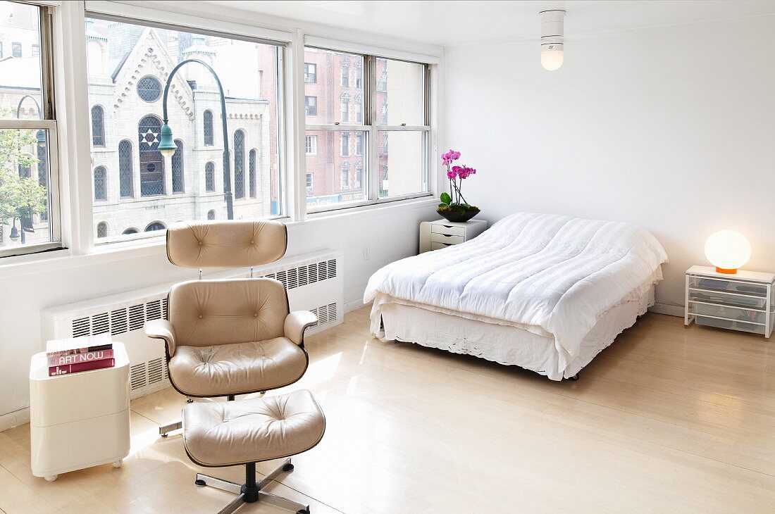 Minimalist bedroom with pale, classic armchair and matching footstool in front of double bed next to panoramic window