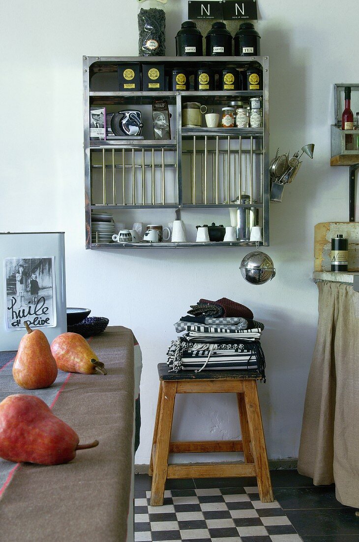 Kitchen utensils, tea caddies and crockery on wall-mounted shelves, tea towels on stool and pears on kitchen table