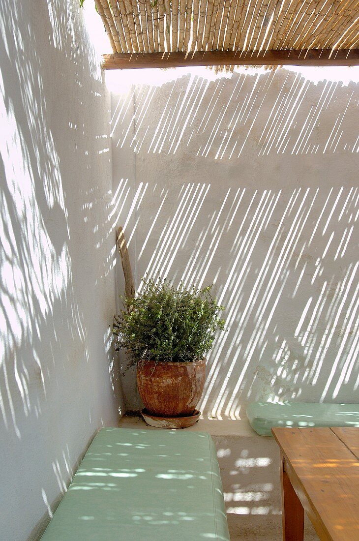 Pale green seat cushions on stone bench in corner against white exterior wall beneath pergola with bamboo roof