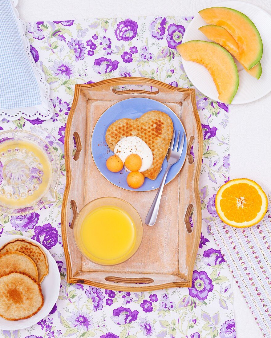 A breakfast tray with a heart-shaped pancake with yoghurt and melon balls and a glass of orange juice