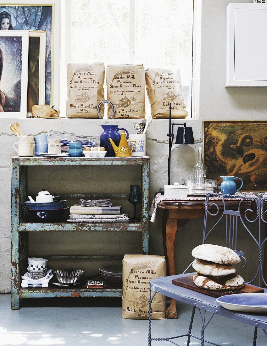 Delicate, blue metal bench in front of vintage shelves with peeling paint against half-height wall and window in workshop-style interior