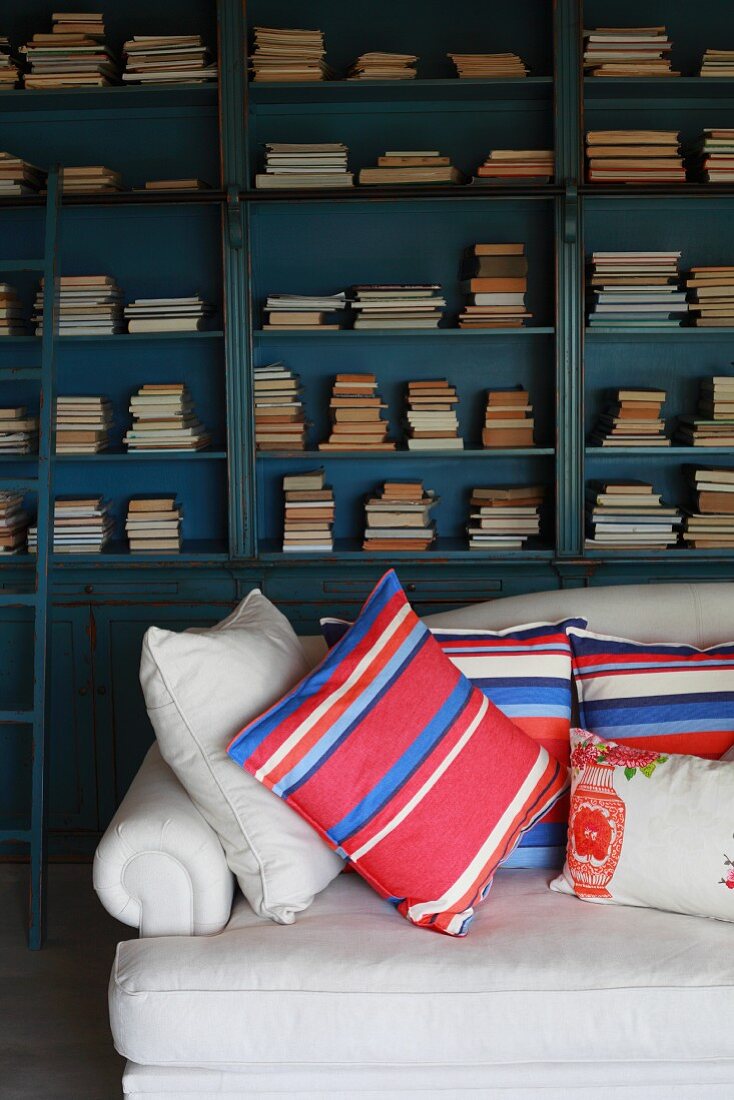 Striped pillows on a sofa upholstered in light fabric in front of a collection of antique books in an open bookcase