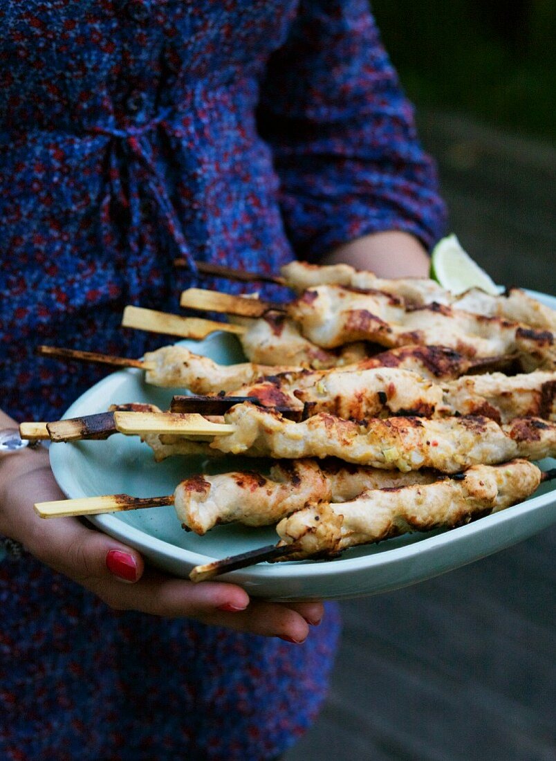 A woman holding a plate of barbecued chicken satay skewers