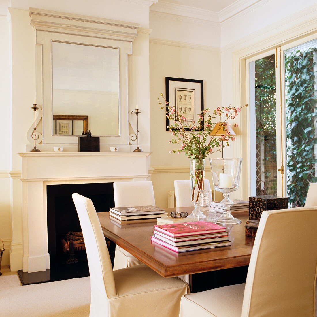 Chairs with pale loose covers and stacked books on wooden table in front of open fireplace in classical living room