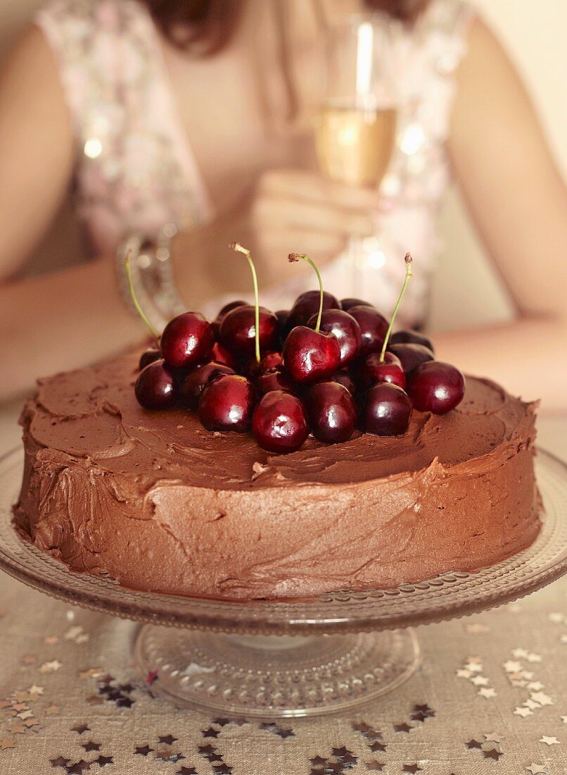 A chocolate layer cake topped with cherries, with a woman holding a glass of sparkling wine in the background
