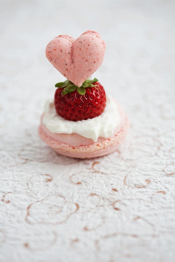 A rose macaroon topped with: cream, a fresh strawberry, and a heart-shaped strawberry macaroon