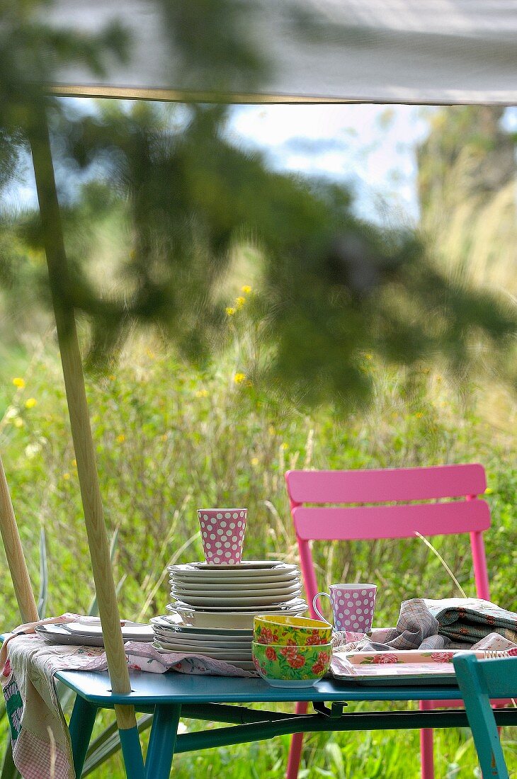 Tellerstapel und bunte Tassen auf einem gedeckten Tisch im Garten unter einem Sonnensegel