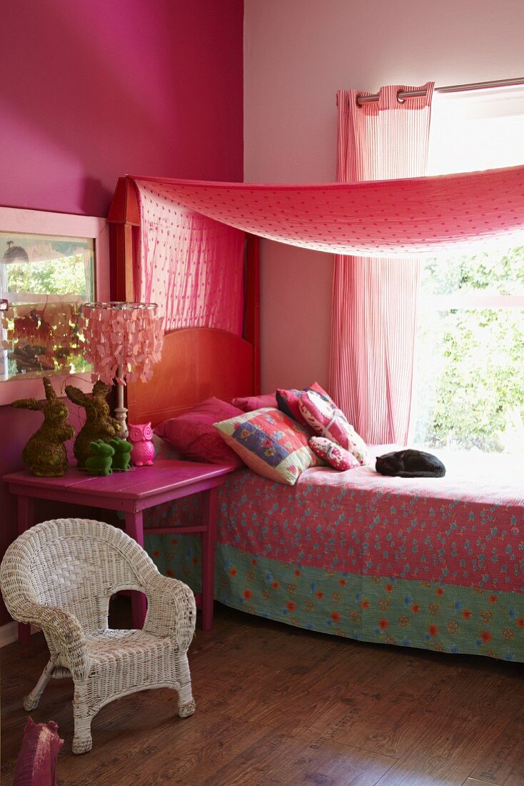 Romantic, girl's bedroom in shades of pink with white wicker chair, pink bedside table, bed with patterned bedspread and canopy