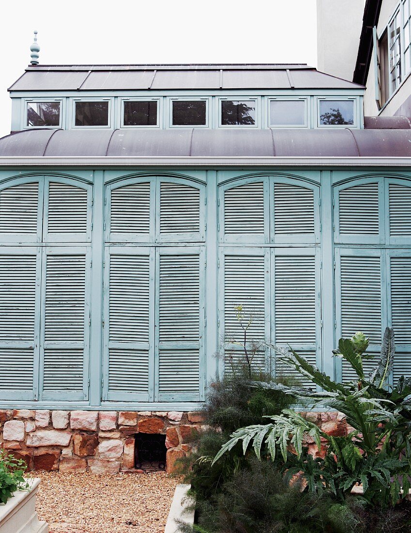Extension with closed, turquoise shutters on stone foundation; raised flower beds and gravel paths