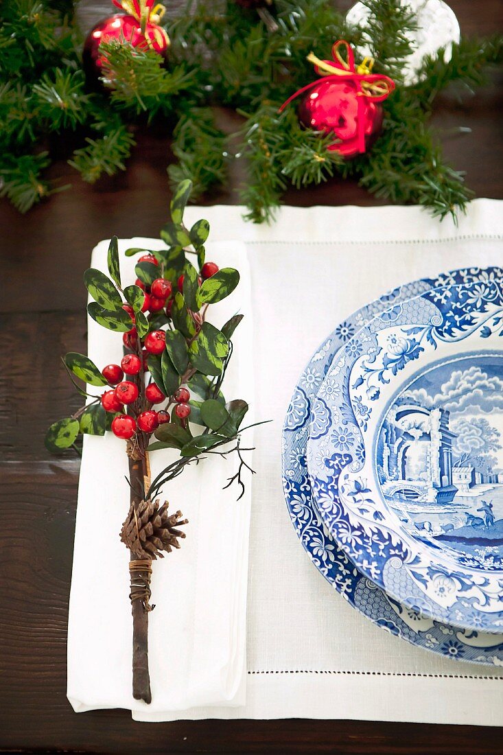 Gedeck mit Blau-weißem Geschirr auf Hohlsaum-Set und kleines Gebinde mit Zapfen und roten Beeren als weihnachtlicher Tischschmuck