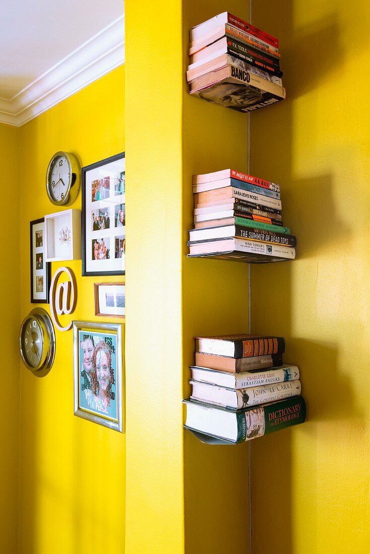 Bilder und Uhren vor gelber Wand, durch Pfeiler von Nische mit Bücherstapeln getrennt