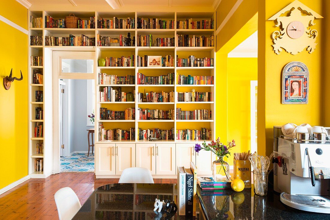 Yellow interior with fitted bookcase; coffee machine on black glossy table and chairs in foreground