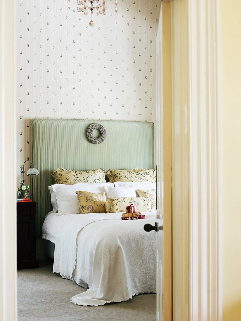 View through open bedroom door of double bed with pastel green, upholstered headboard decorated with silver wreath