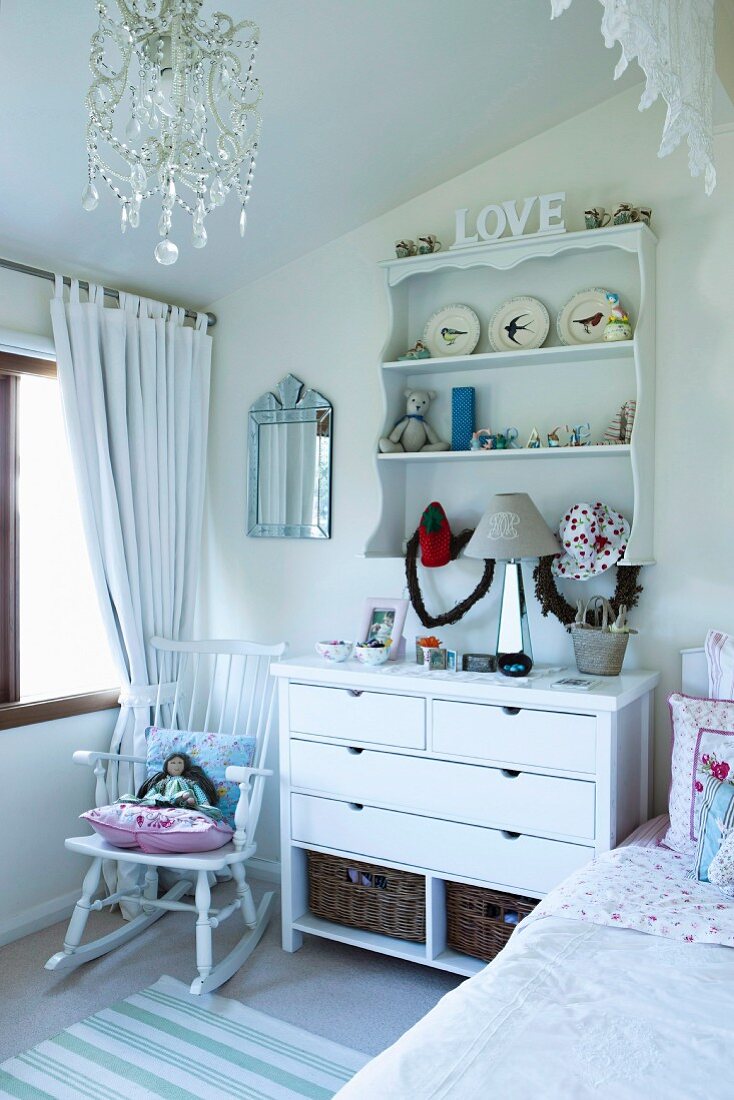 Romantic ornaments on chest of drawers and wall-mounted shelves and doll on rocking chair in white, child's bedroom