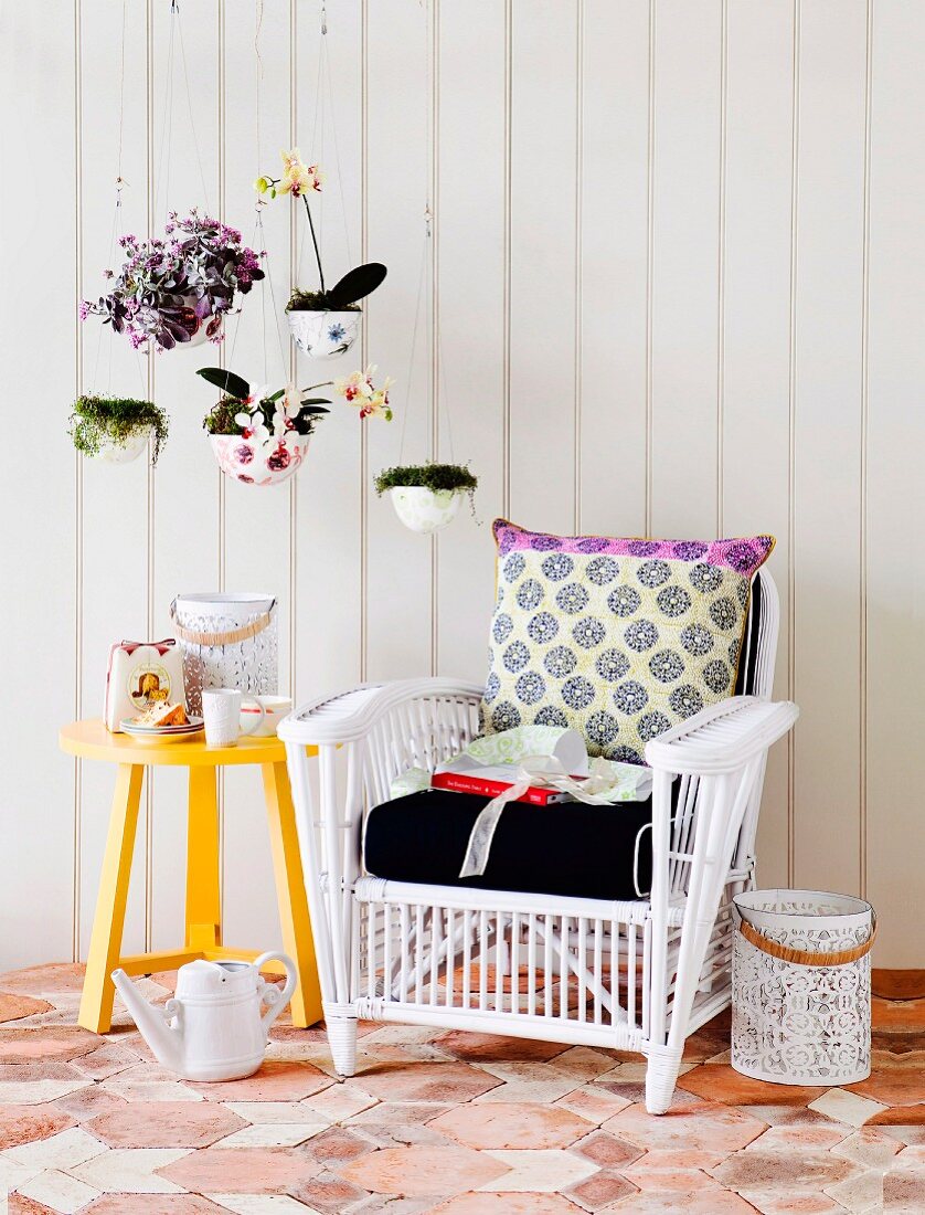 White wicker chair with cushions next to yellow-painted side table against white wooden wall and house plants in planters suspended from ceiling