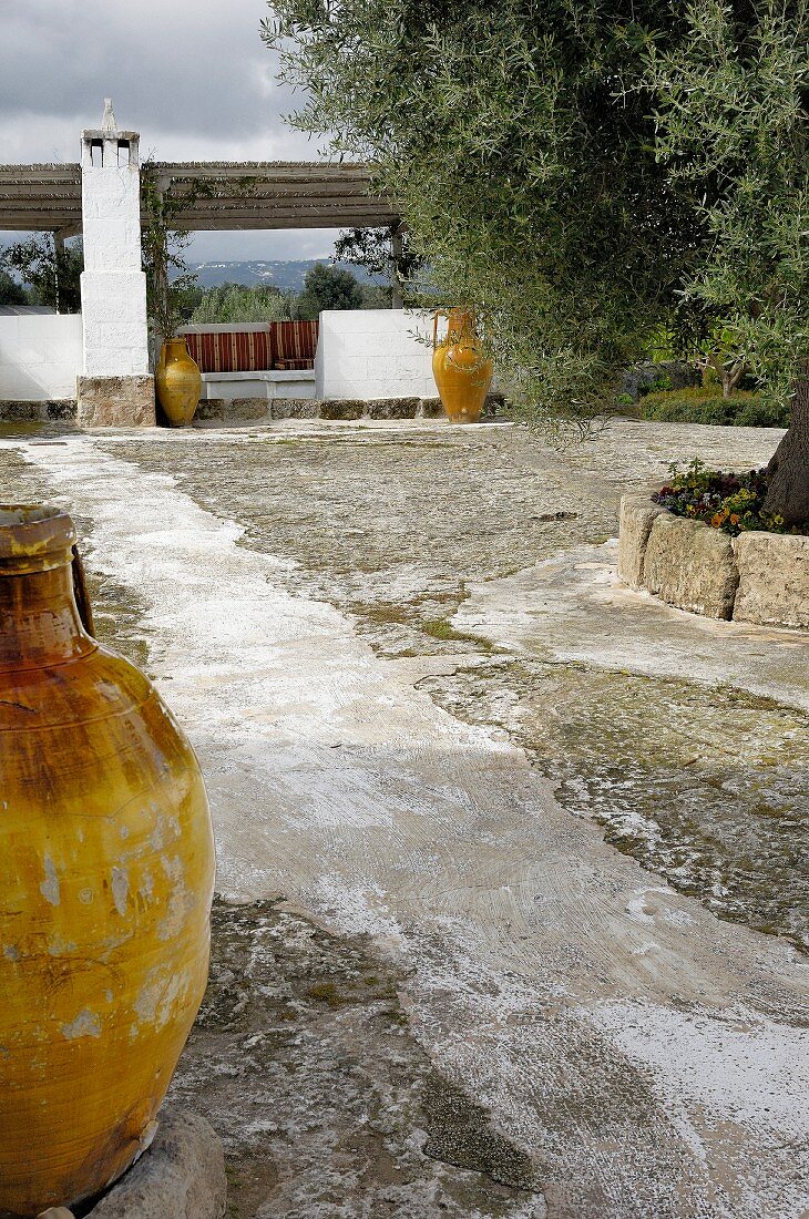Rustic courtyard with yellow amphorae; pergola with bench in background