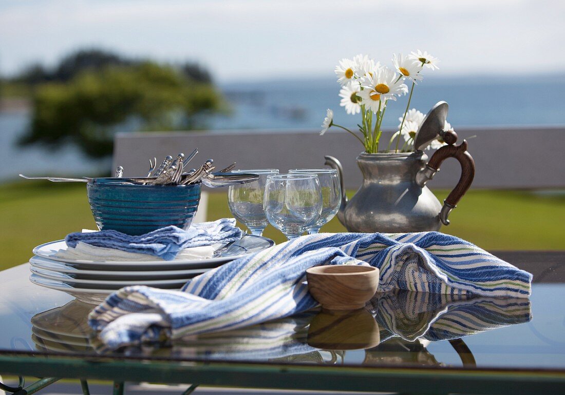 Gedeckter Tisch am Meer mit Geschirr, Weingläsern, Zinnkrug mit Blumen und Tuch