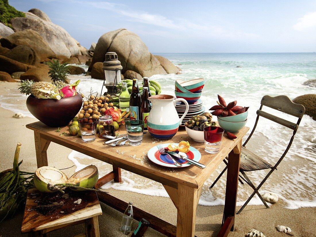 Tisch mit exotischen Früchten, Getränken und Geschirr am Meer