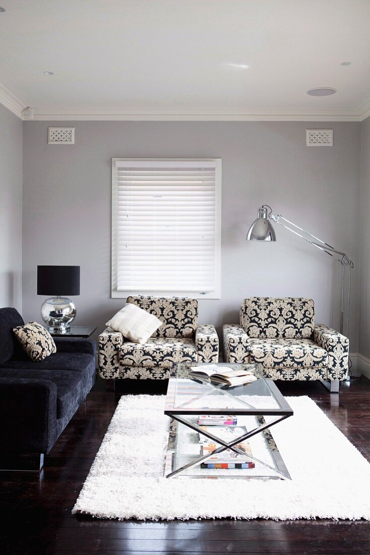 Glass table with metal frame on pale flokati rug in front of patterned armchairs and sofa in traditional living room with walls painted pale grey