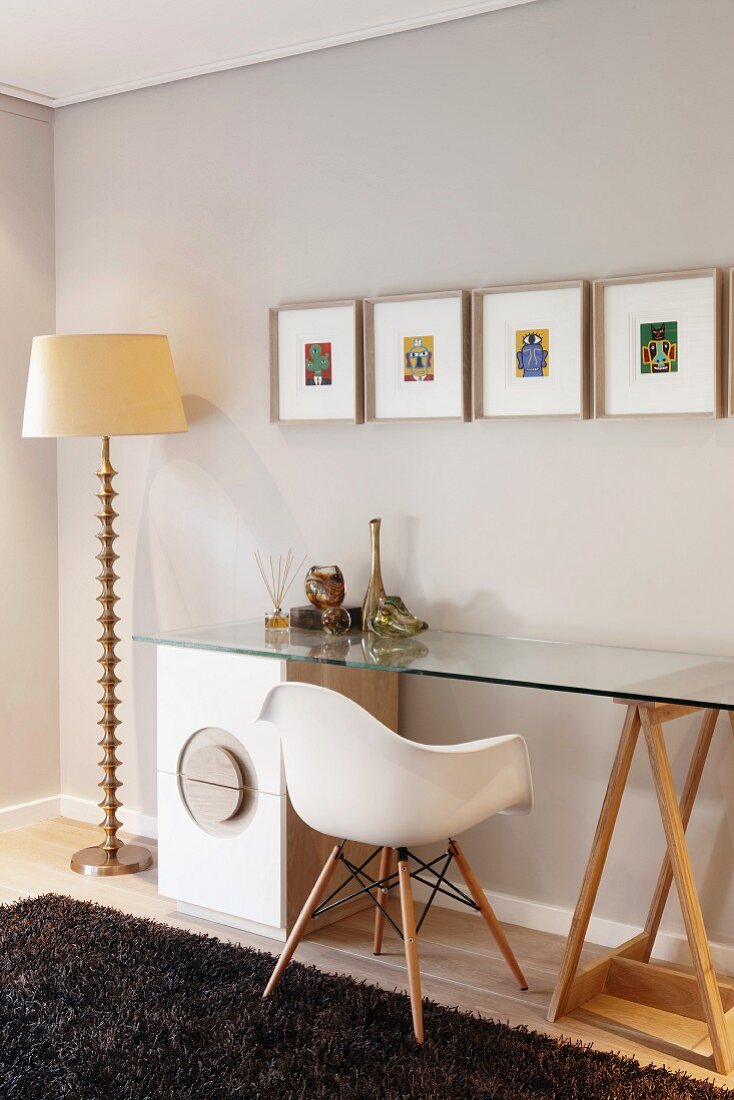 Classic chair at glass table top resting on trestle and drawer cabinet; standard lamp with ornate metal base and small, colourful artworks on wall