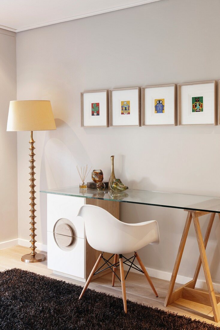 Klassikerstuhl an einem Glastisch mit Schubladenschrank; Stehlampe mit verziertem Metallfuss und bunte Miniaturbilder an der Wand