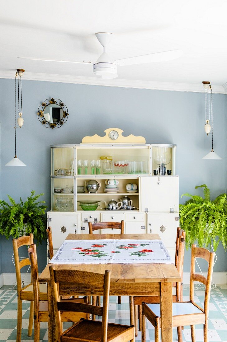 Kitchen-dining room with simple, plain wood dining set; antique kitchen dresser, adjustable pendant lamps and ferns against pale blue wall