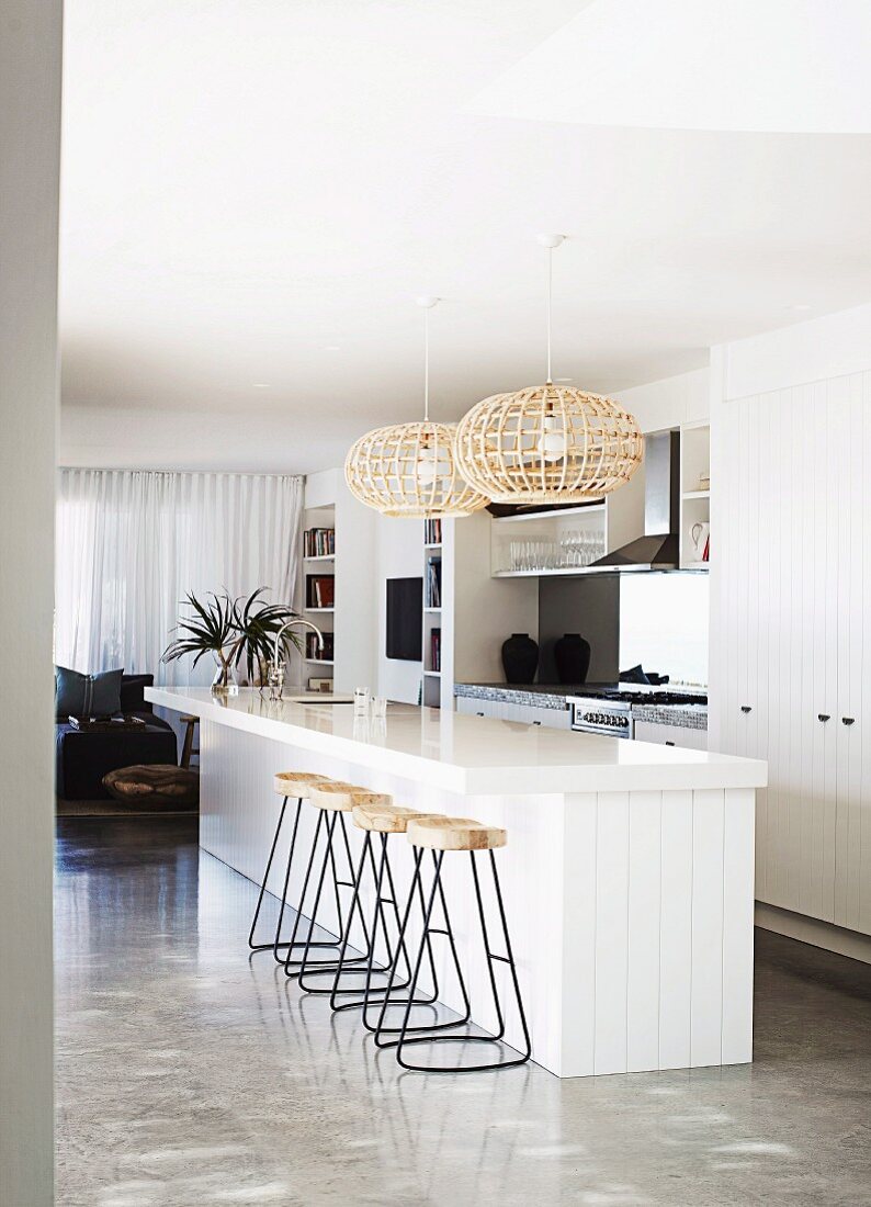 Barstools with metal frames at white, free-standing kitchen counter below wicker pendant lamps in designer apartment