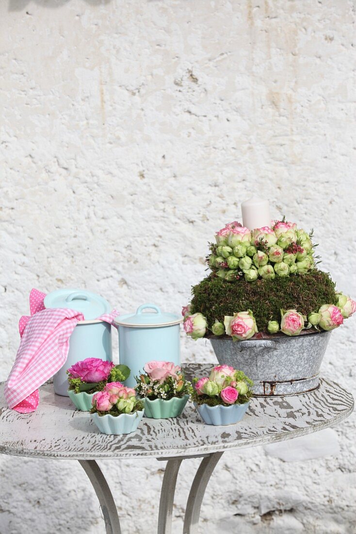 Flower arrangements with pink roses, moss and candles in old enamel pan next to rose posies in china flan dishes