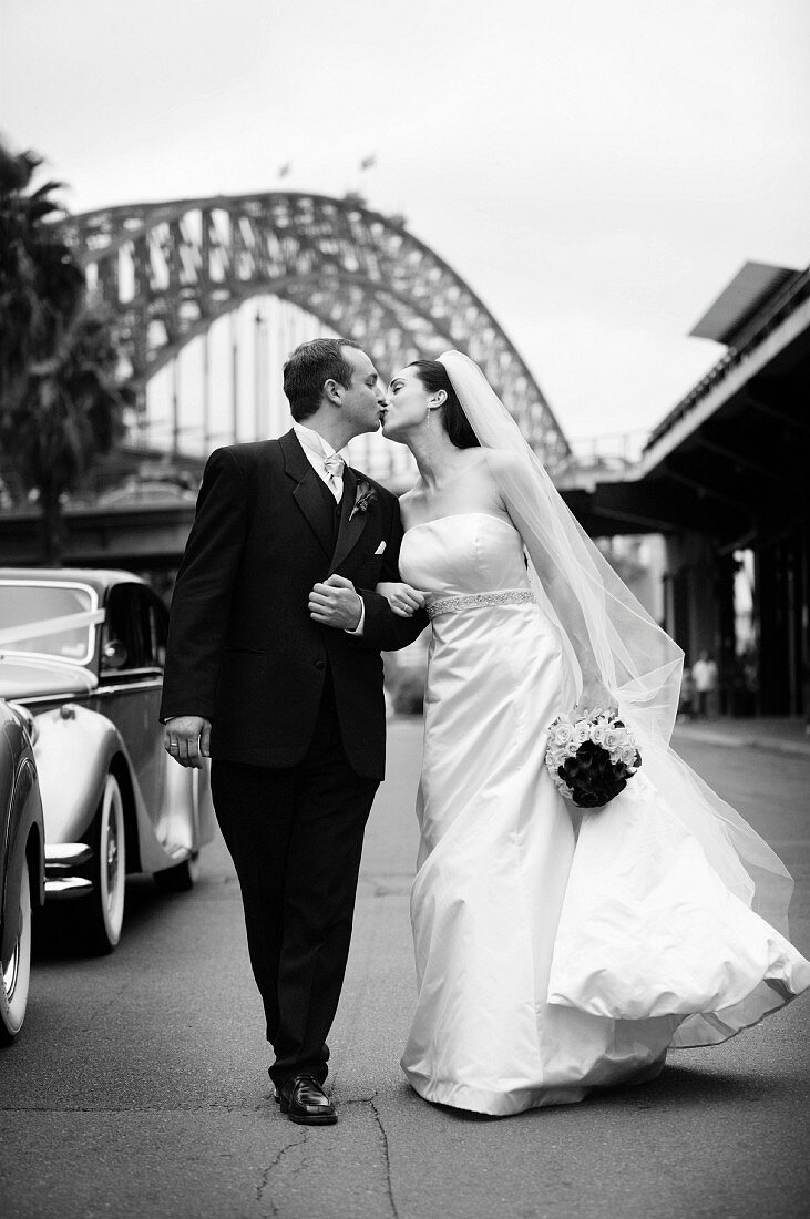 Bride wearing long white dress and groom in black suit kissing whilst walking down the street (black and white photo)