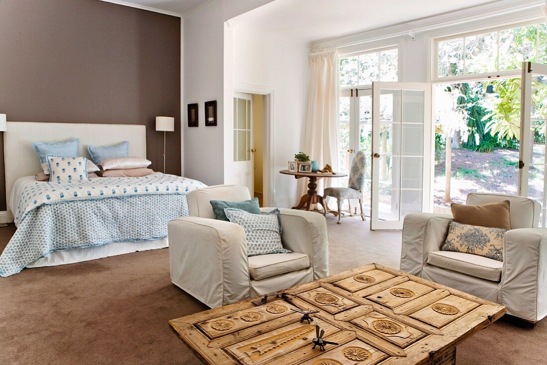 Offener Raum mit Hussensesseln und geschnitztem Türblatt-Tisch; im Hintergrund Doppelbett mit pastellfarbener Bettwäsche