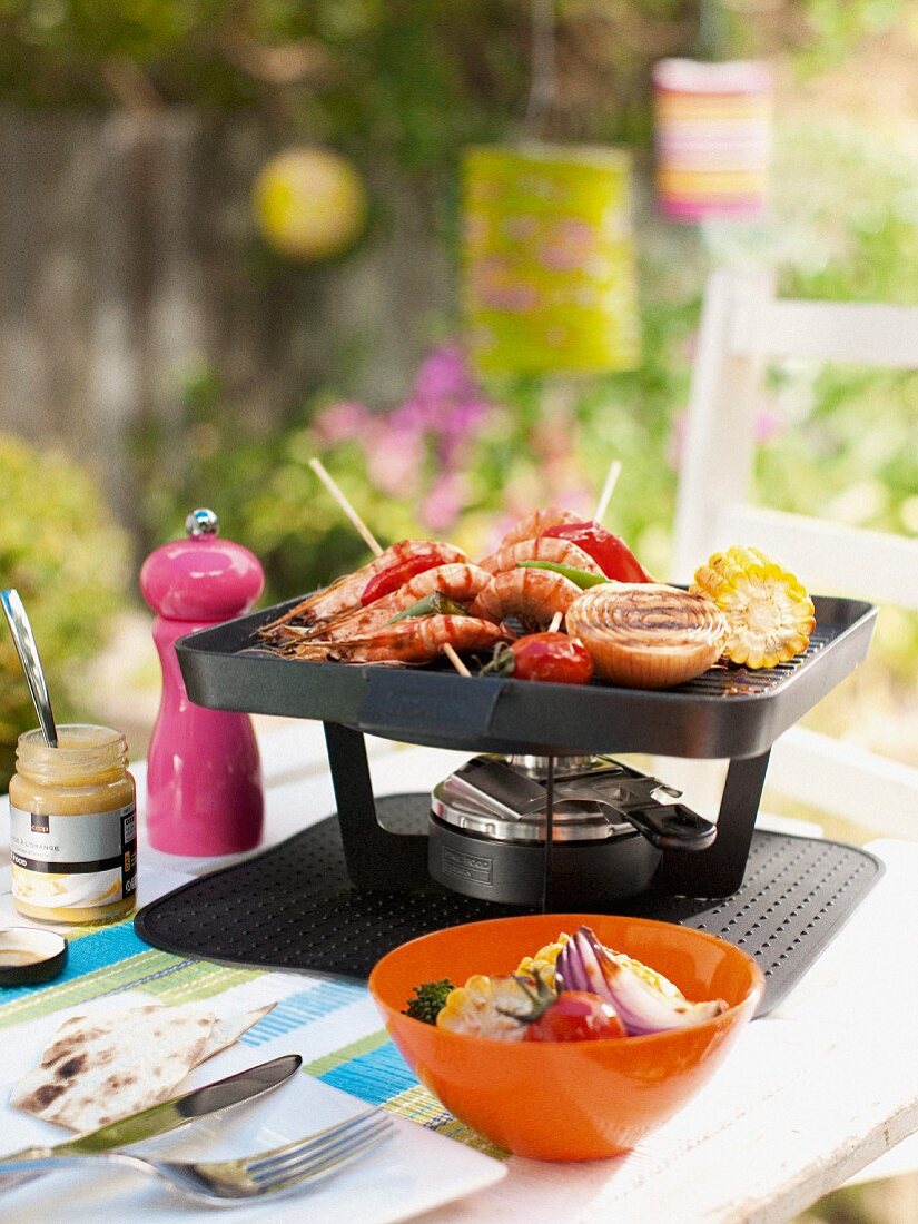 A table-top barbecue with prawn skewers and vegetables, on a garden table