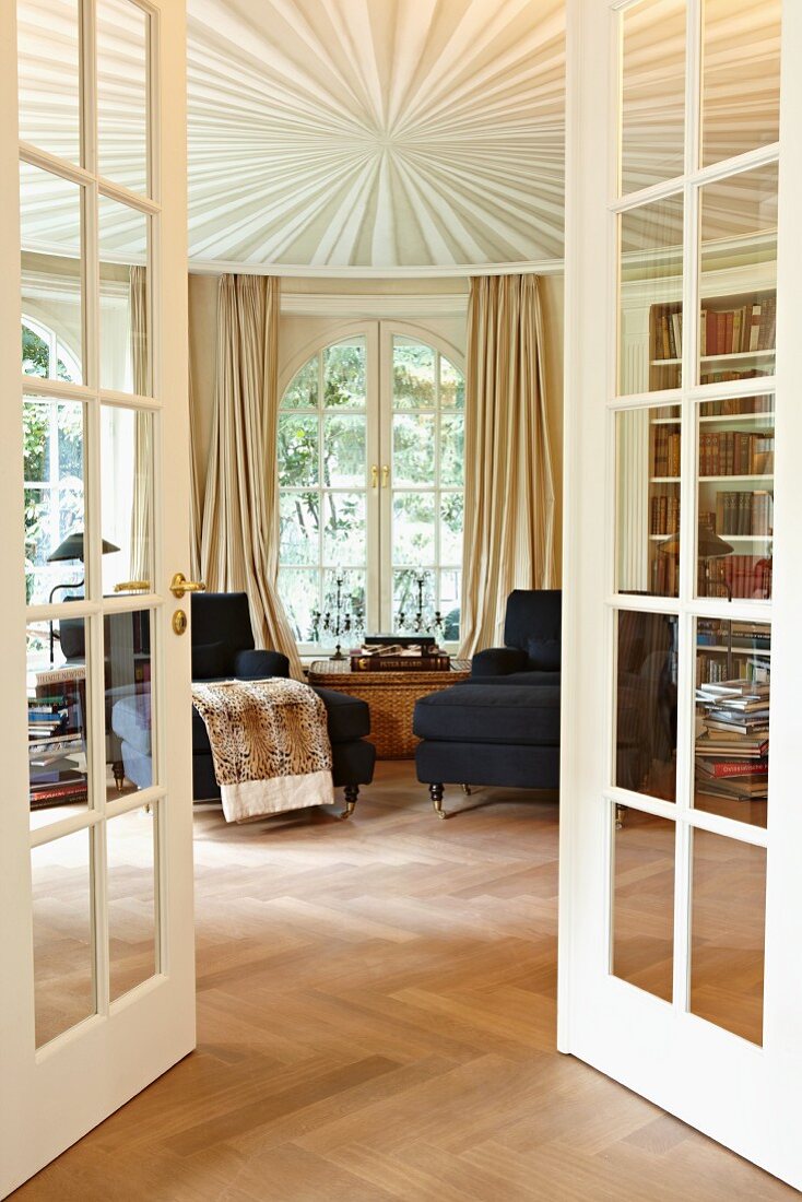View through open French doors of a chaise lounge by a window and a suspended ceiling with a star-shaped design in the circular drawing room of a stately home
