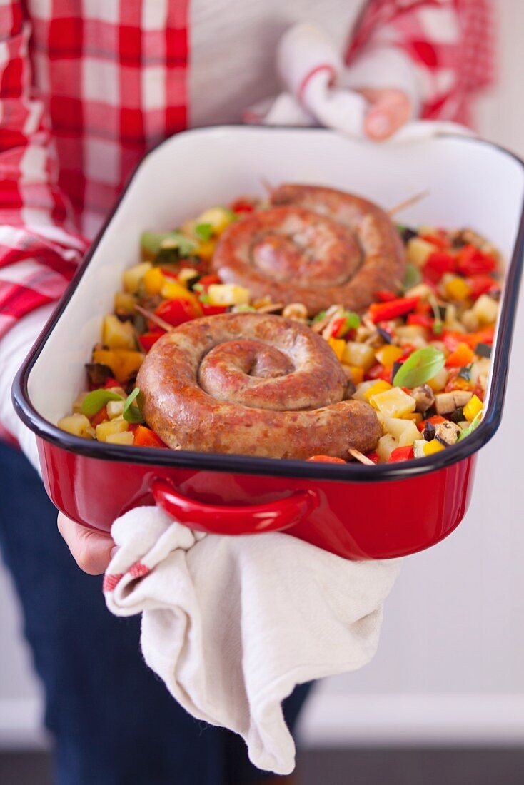 Woman holding baking dish with spiral pork sausages on baked vegetables