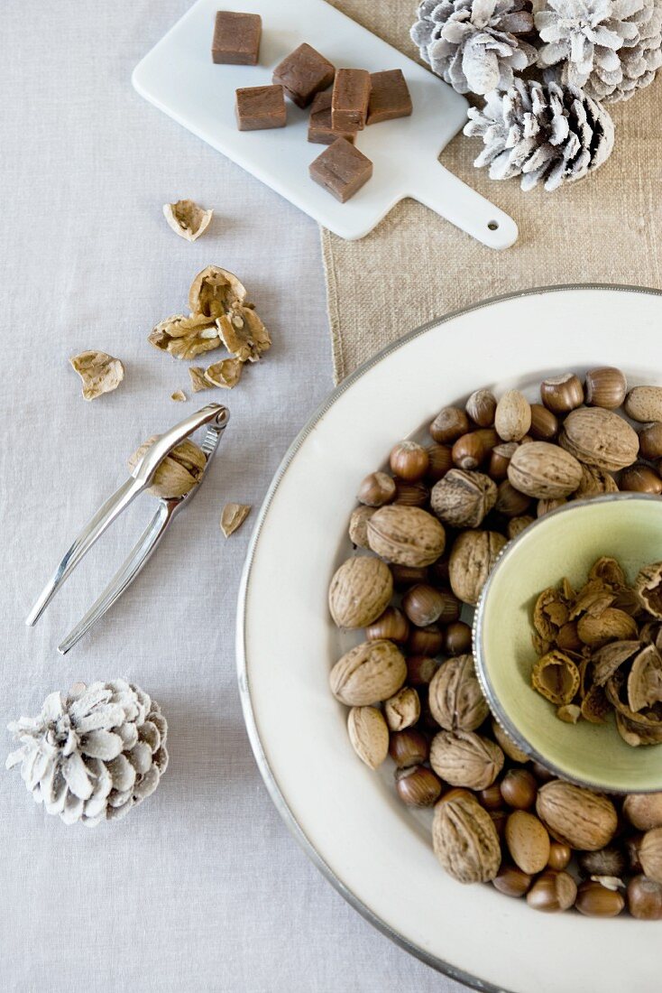 Weihnachtliche Tischdeko in Naturfarben mit Nüssen auf hellen Tellern mit Silberrand, Schokostückchen auf Porzellanbrett und weissen Tannenzapfen