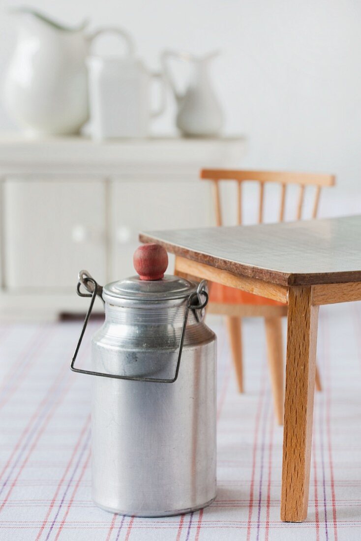 Still-life of dolls' house furniture: milk can, table, chair, sideboard and jugs on checked cloth