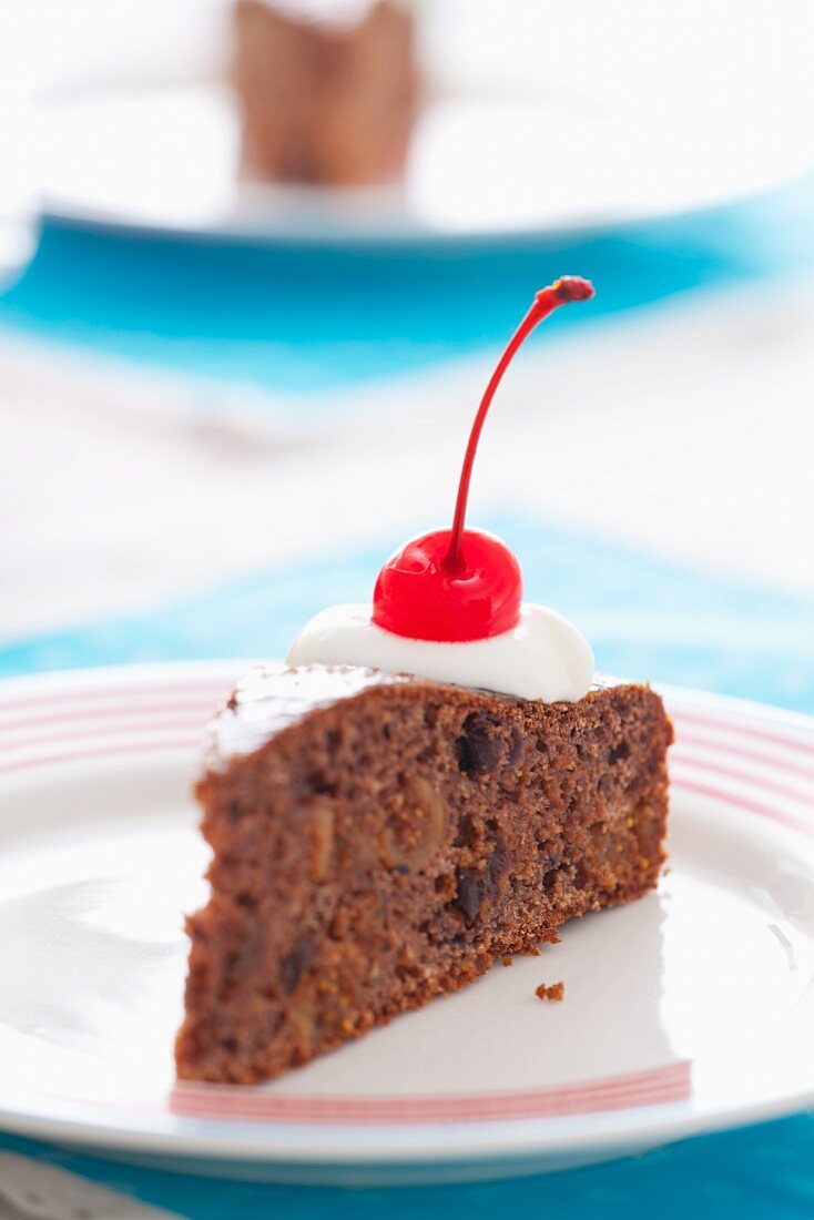 A slice of chocolate cake with dried fruit, cream and a glacé cherry