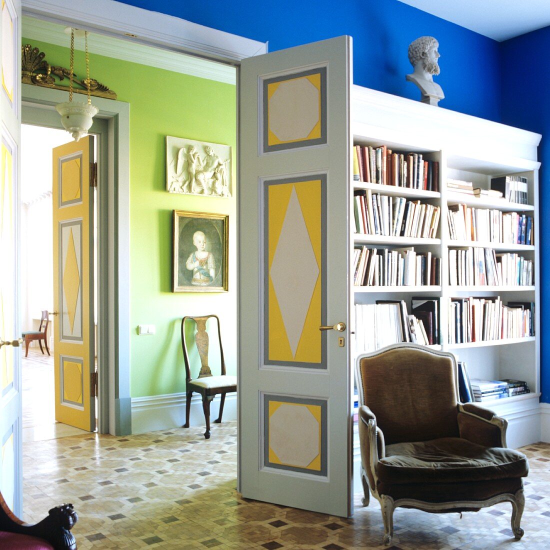 Rococo armchair in front of fitted bookshelves in blue interior; view into green room through open double doors in grand house