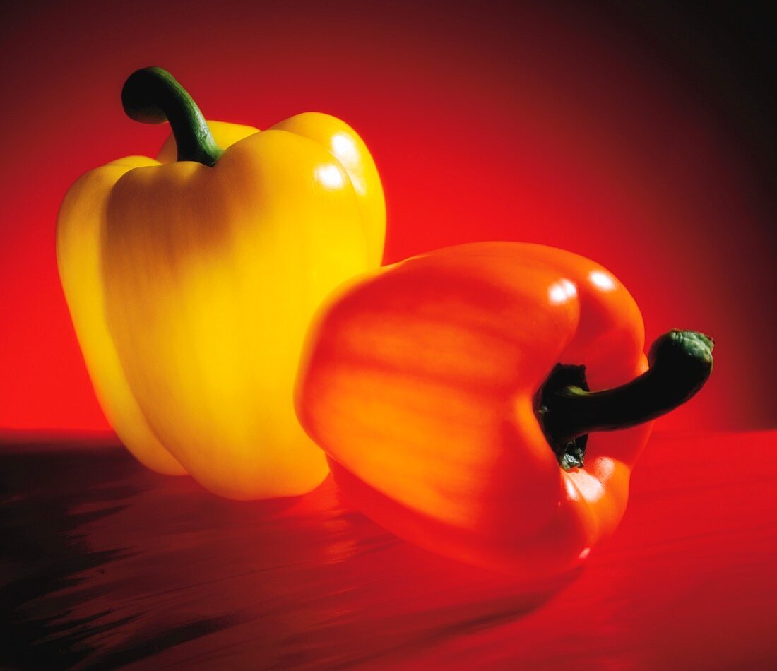 An orange and a yellow pepper on a red surface