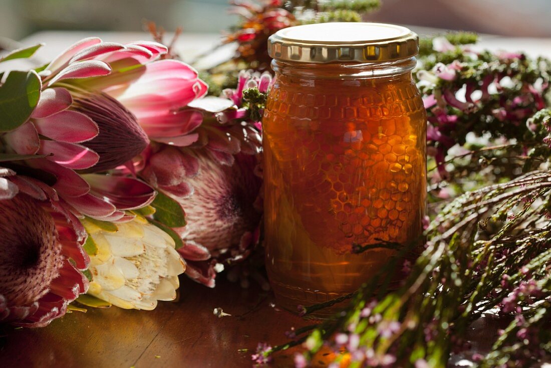 A jar of honey with honeycomb, surrounded by flowers