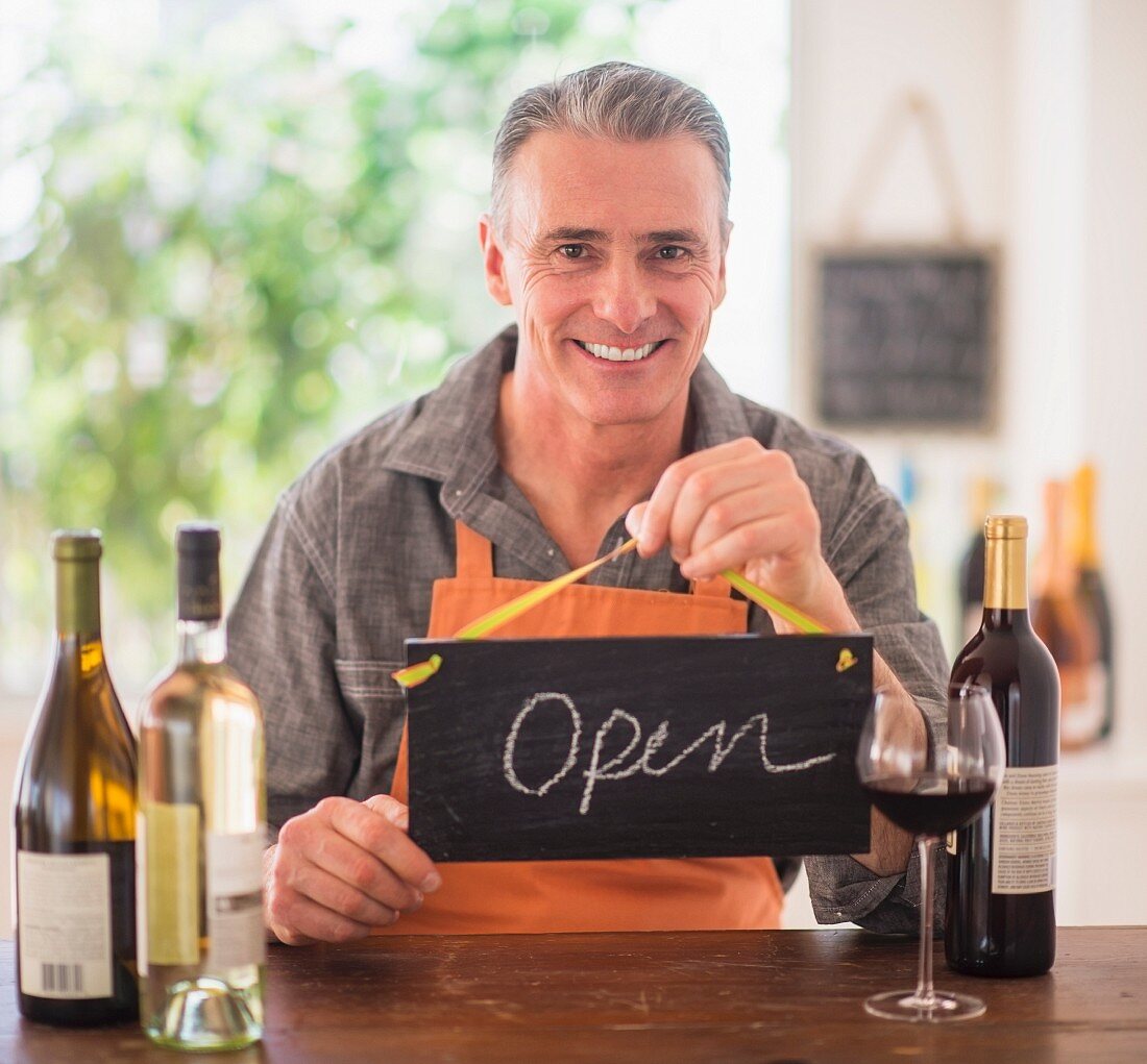 A man in a wine shop holding a sign saying 'Open'