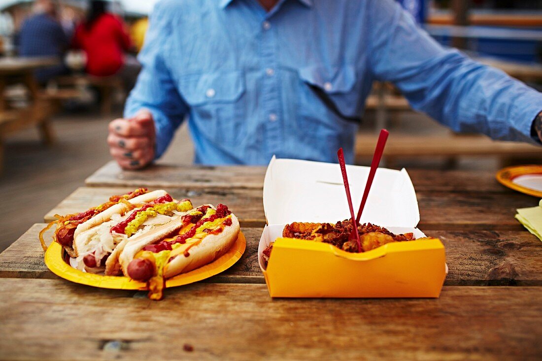 Mann sitzt an Holztisch vor Fast Food & Hot Dogs