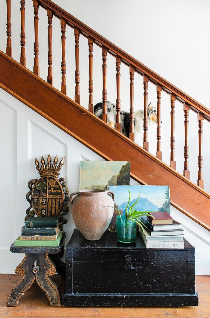 Antiquarian books on black, wooden trunk and footstool in front of staircase with wooden balustrade