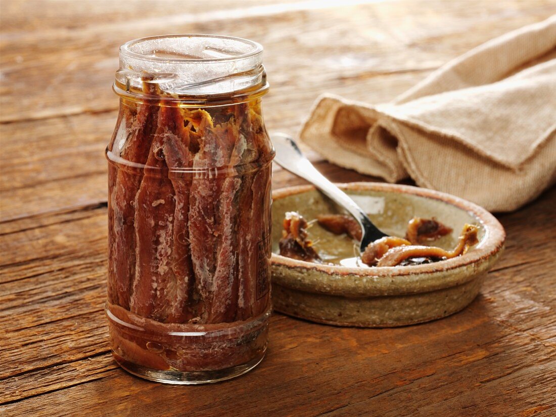 Jar of anchovies on old barnwood with a few in ceramic dish with a fork